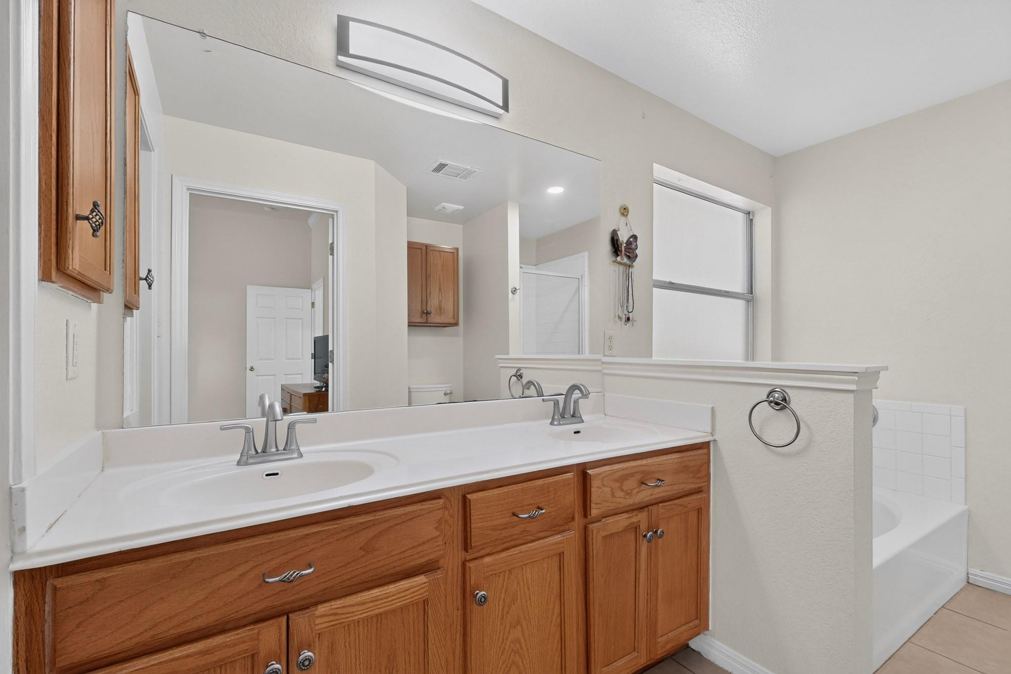 Bathroom with a double-sink wood vanity, large mirror, and bathtub beside a window.