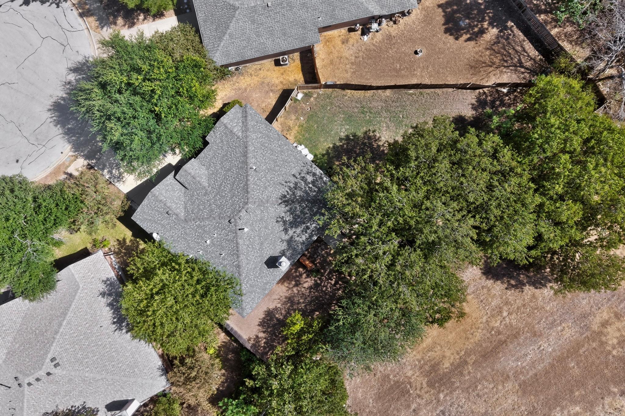 Aerial view of a gray-roofed home surrounded by mature trees, with a fenced backyard and paved patio.