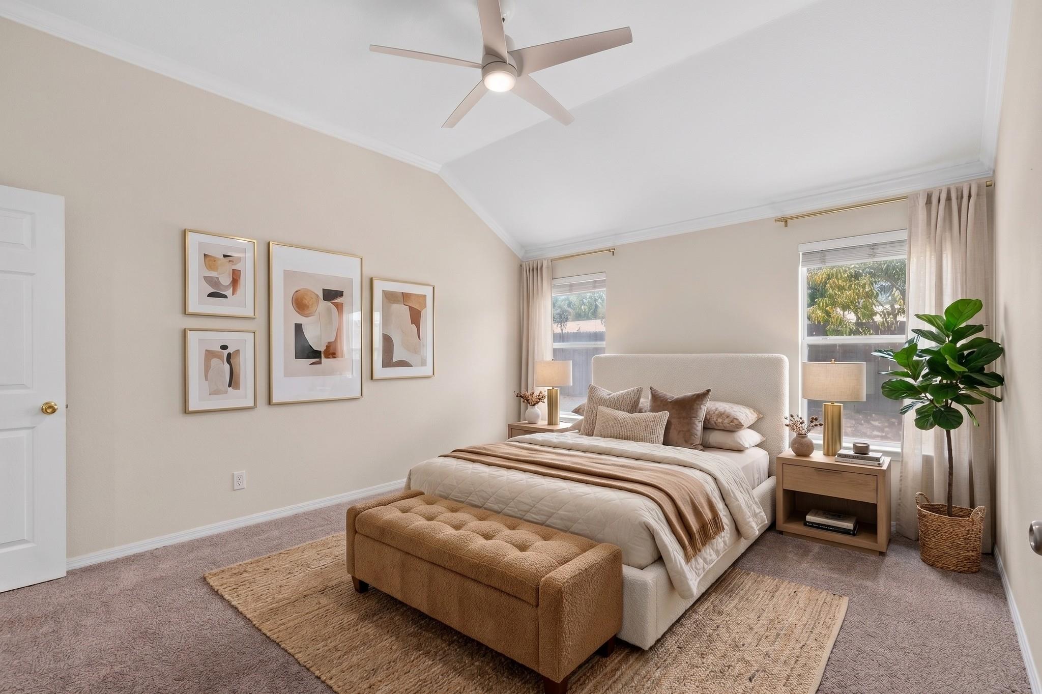 Staged bedroom with a king-size bed, tan tufted bench, framed art, and large curtained windows.