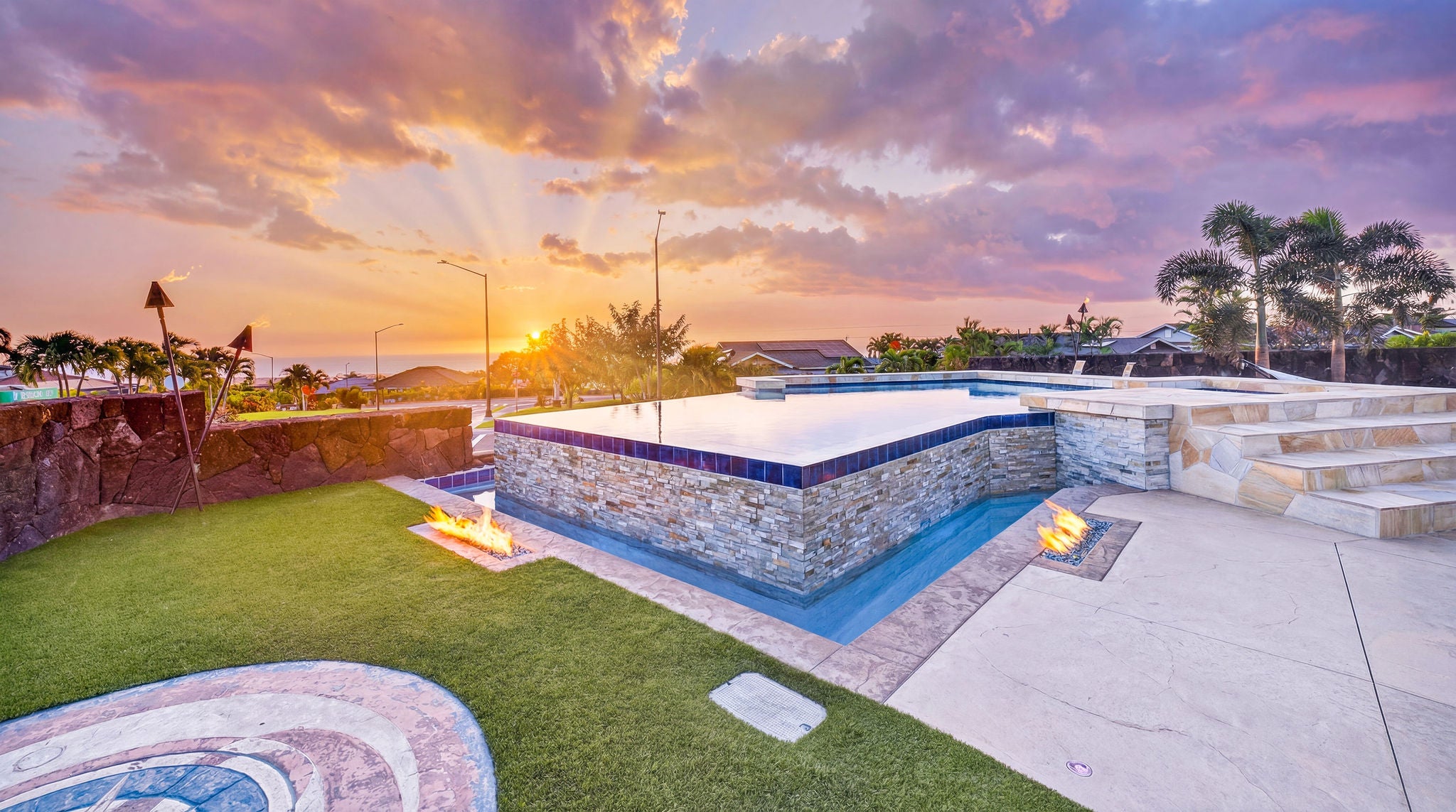 Stone-sided pool and patio with fire features, palm trees, and an ocean view at sunset.