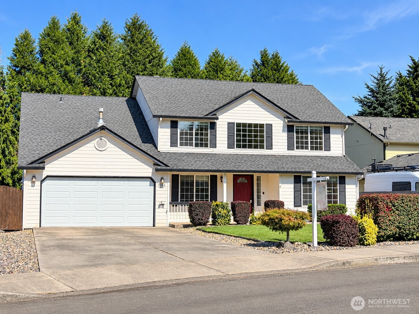 Two-story house with a red front door, double garage, landscaped yard, and tall evergreens behind it.
