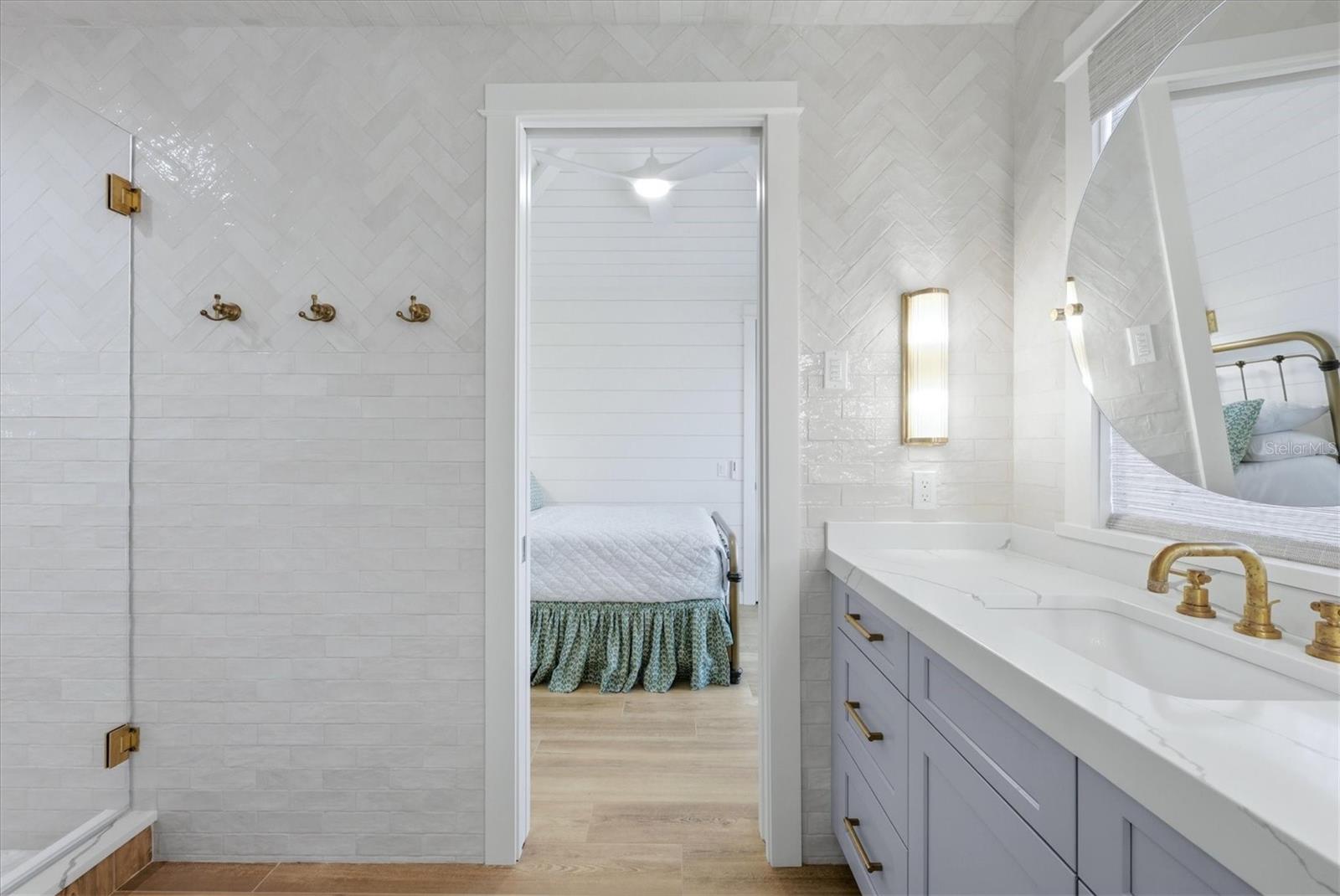 Bright tiled bathroom with brass fixtures and a lavender double vanity, opening onto a bedroom.