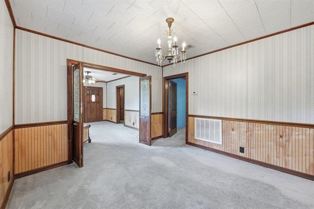 Vacant room with gray carpet, wood wainscoting, and a chandelier, opening into adjoining rooms.