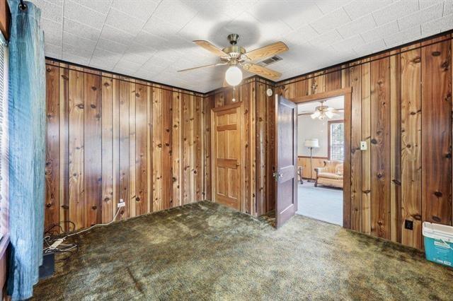 Wood-paneled interior room with mottled carpet and a ceiling fan, looking through an open doorway to a furnished room.