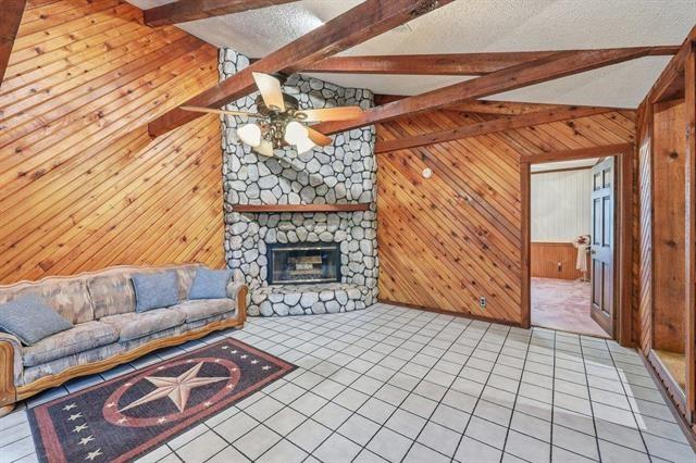 Living room with a stone fireplace, vaulted wood ceiling, exposed beams, and tiled floor.