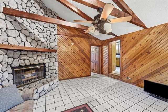 Living room with a stone fireplace, wood-paneled walls, exposed beams, and tiled flooring.