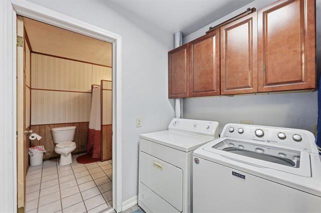 Laundry room with white washer and dryer beneath wood cabinets, beside a bathroom doorway.