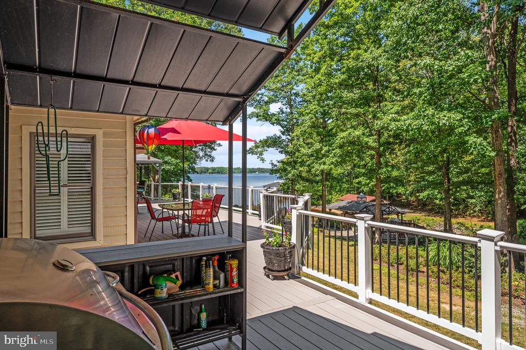 Covered deck with a grill and red umbrella overlooking a lake and wooded shoreline.