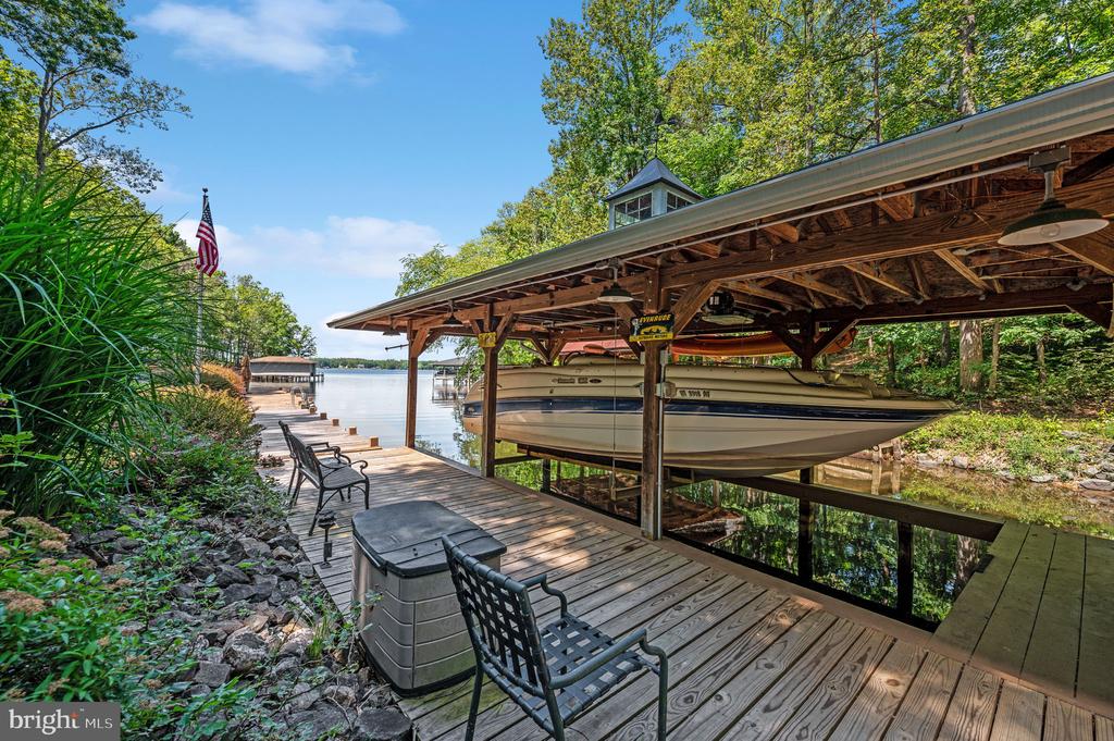 Waterfront dock with a covered boat lift, motorboat, hot tub, and seating beside a tree-lined lake.