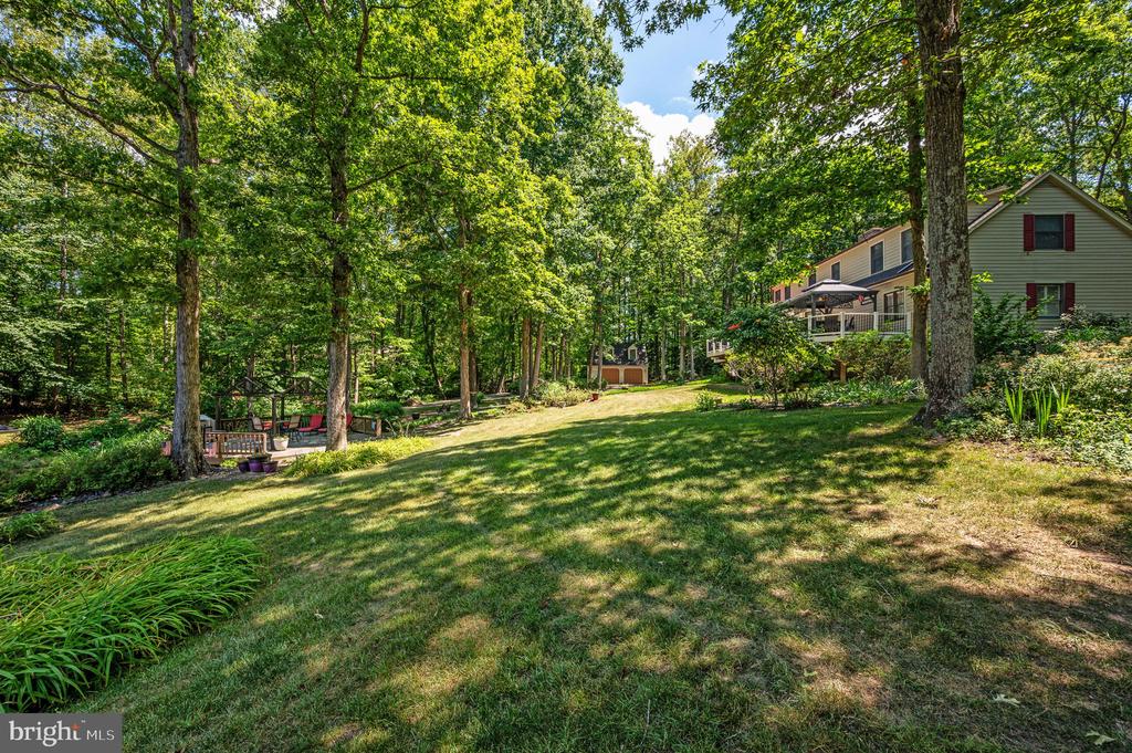 Wooded backyard with a sloping lawn, landscaped beds, patio seating, and the house at right.