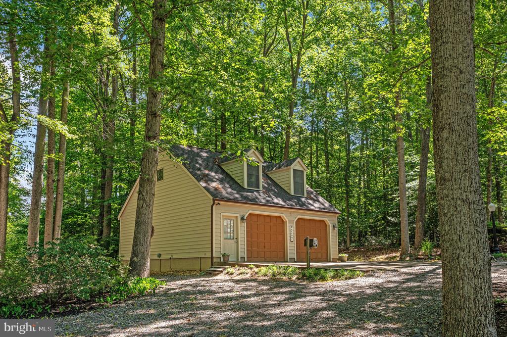 Detached two-car garage with dormers, set among leafy trees beside a gravel driveway.