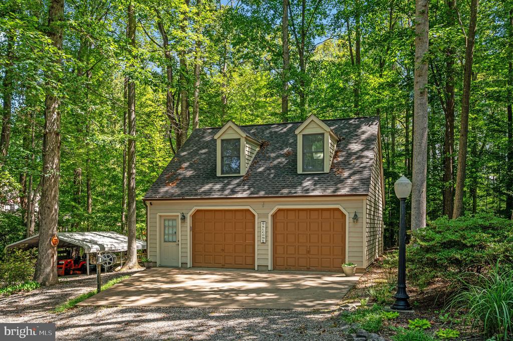 Detached two-car garage with dormer windows, surrounded by mature trees; a covered boat shelter stands beside the driveway.