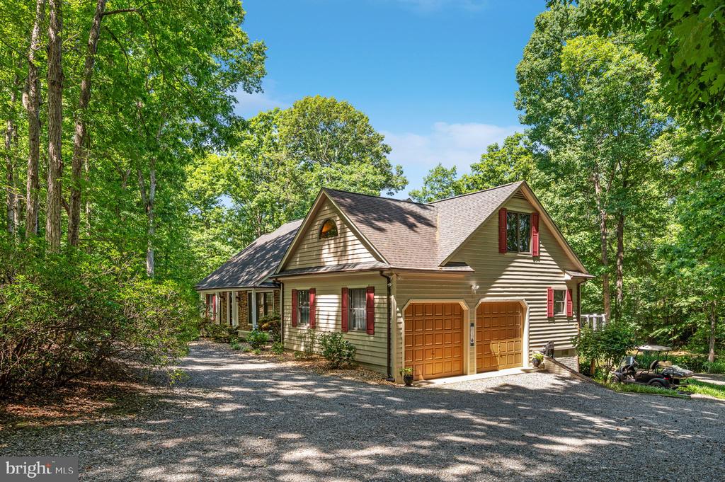 Tan house with red shutters and double garage doors, surrounded by trees beside a gravel driveway.