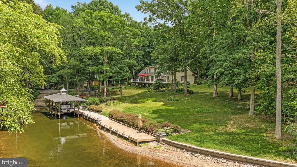Waterfront house above a dock and covered boat slip, surrounded by a wooded lawn.