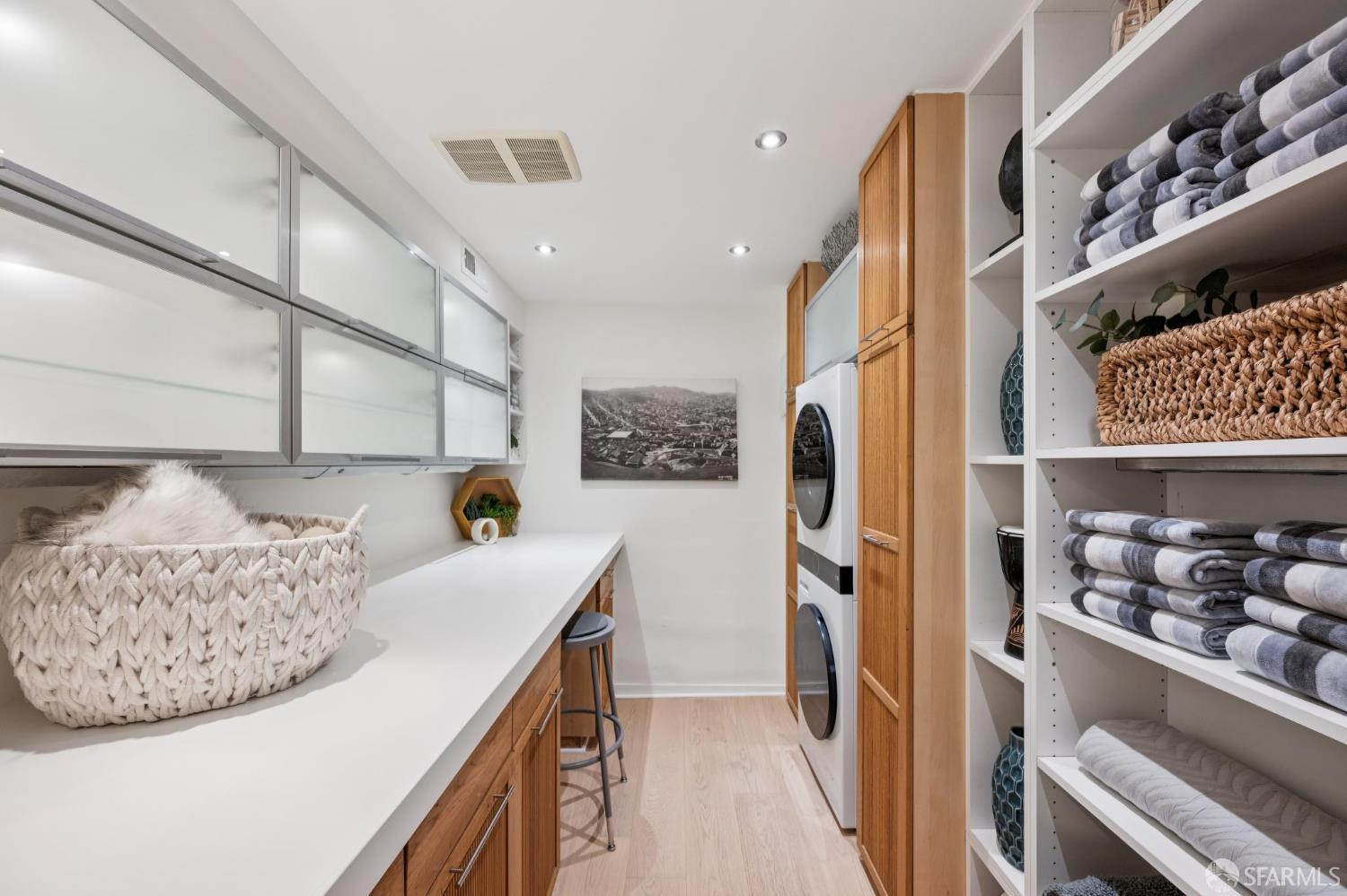 Laundry room with stacked washer and dryer, long counter, wood cabinets, and towel-filled shelving.