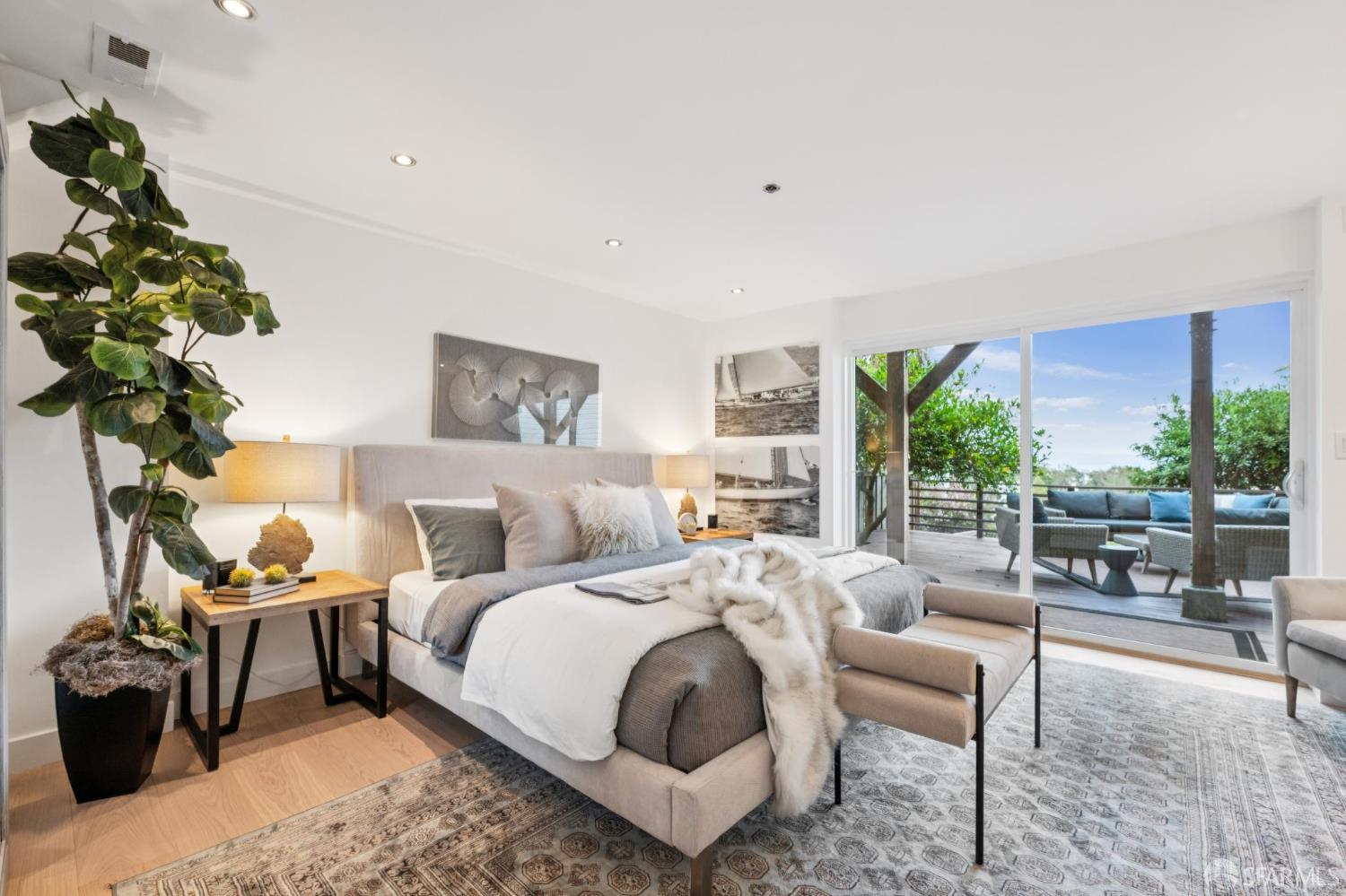 Staged bedroom with a large upholstered bed and sliding doors opening onto a leafy, furnished deck.