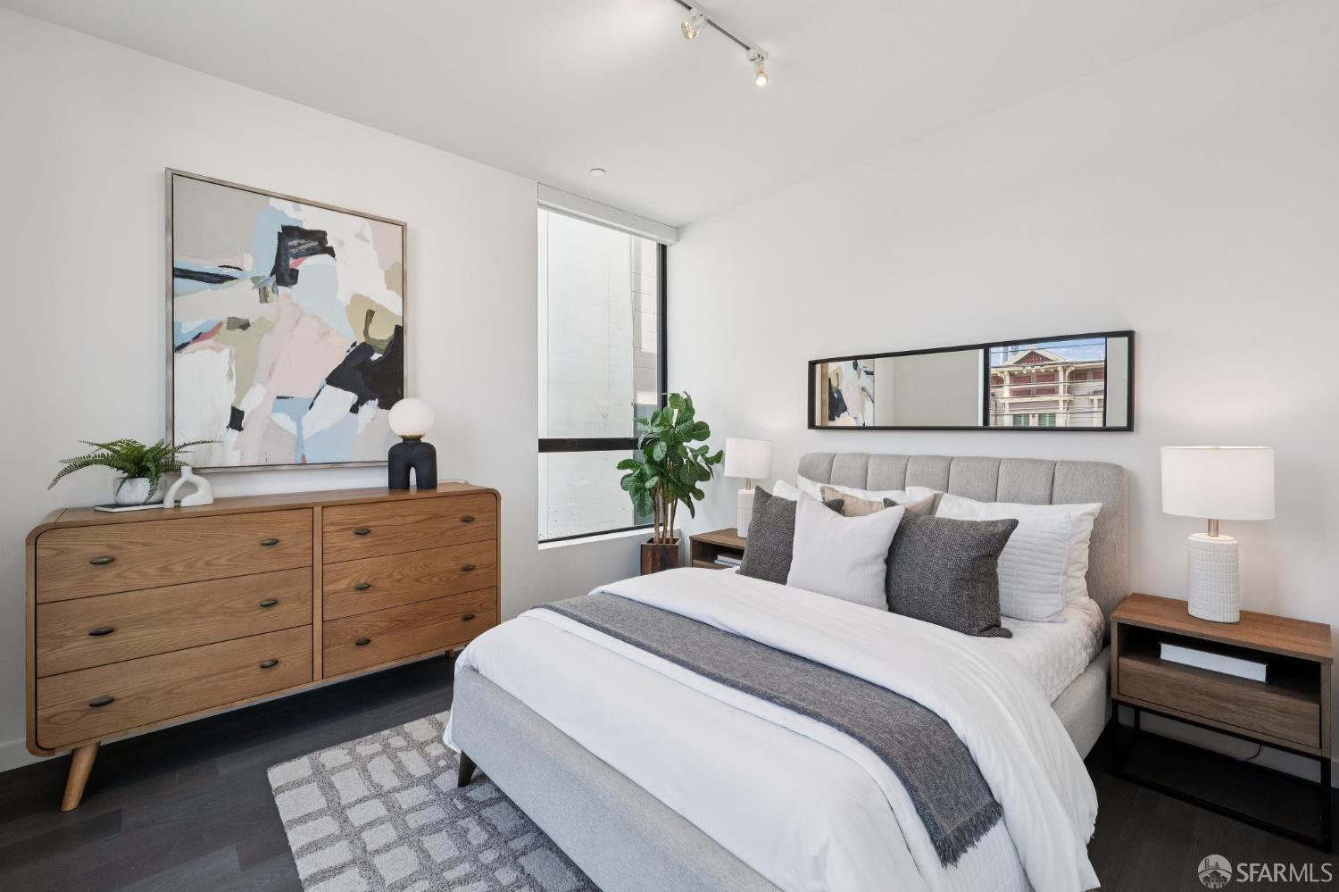 Staged bedroom with a white bed, gray accents, wood dresser, abstract art, and tall window.