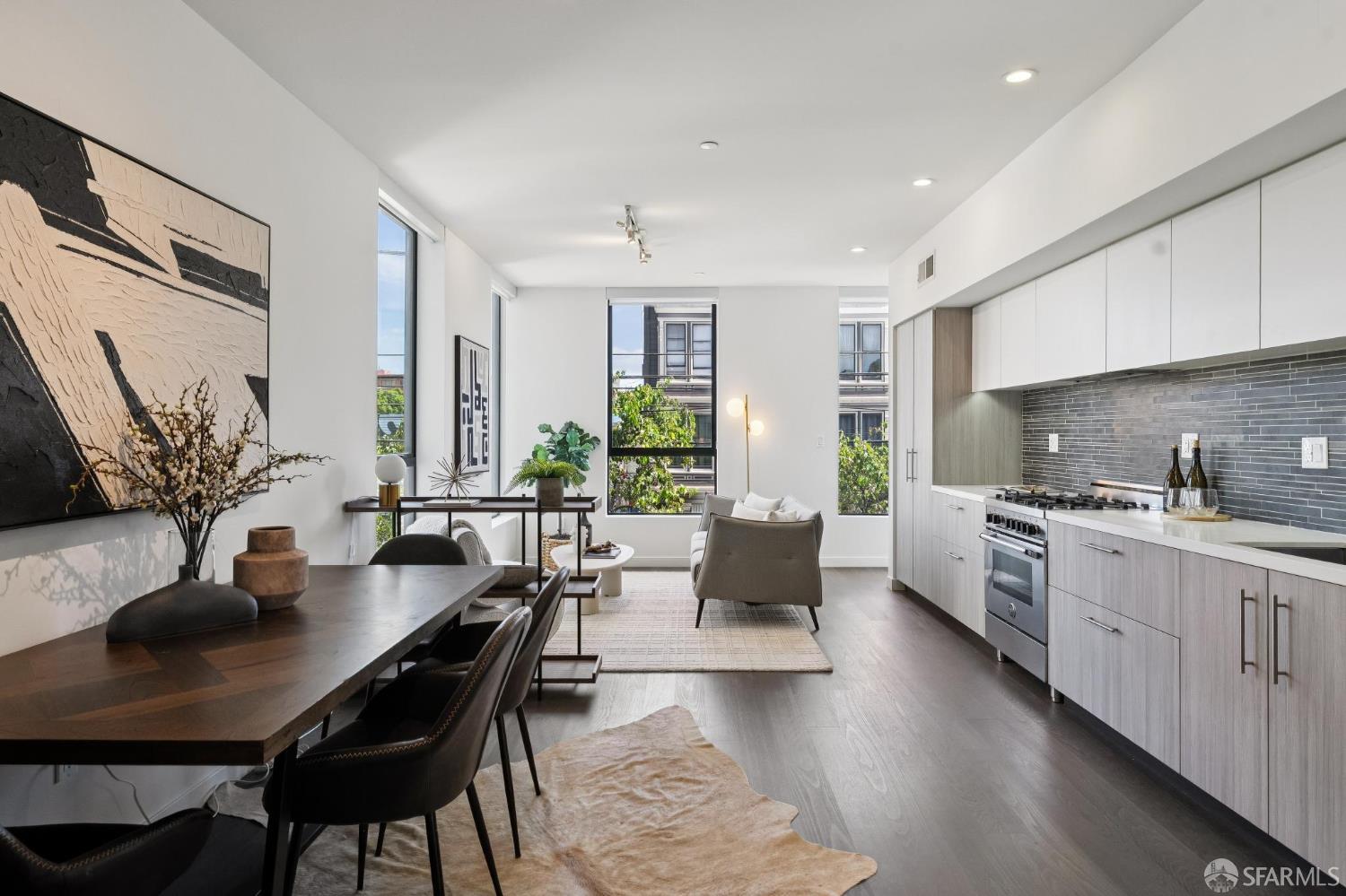 Staged open-plan kitchen and dining area with gray cabinetry, dark tile backsplash, and tall windows.