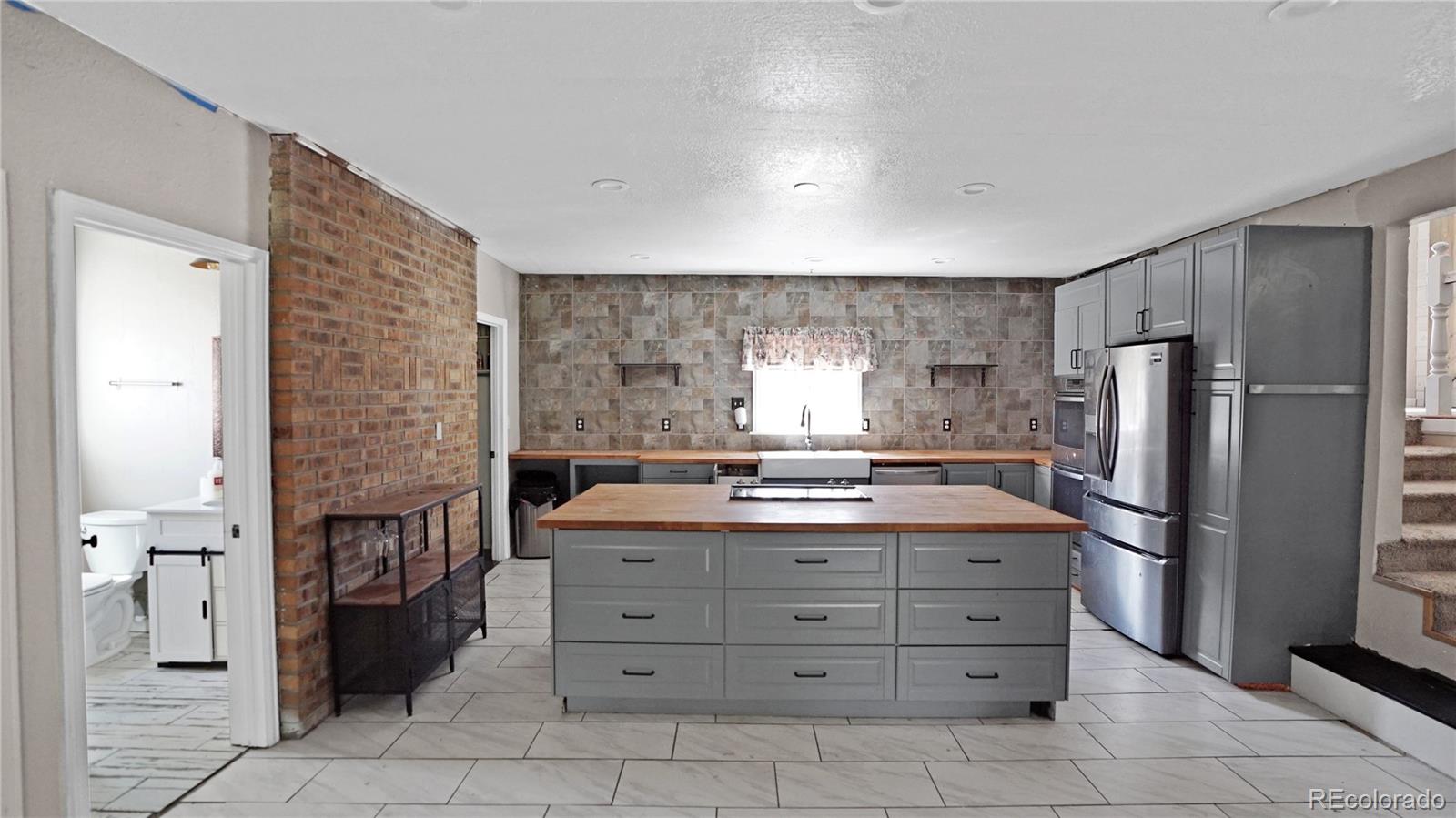 Kitchen with a wood-topped island, gray cabinets, stainless steel appliances, and a brick accent wall.
