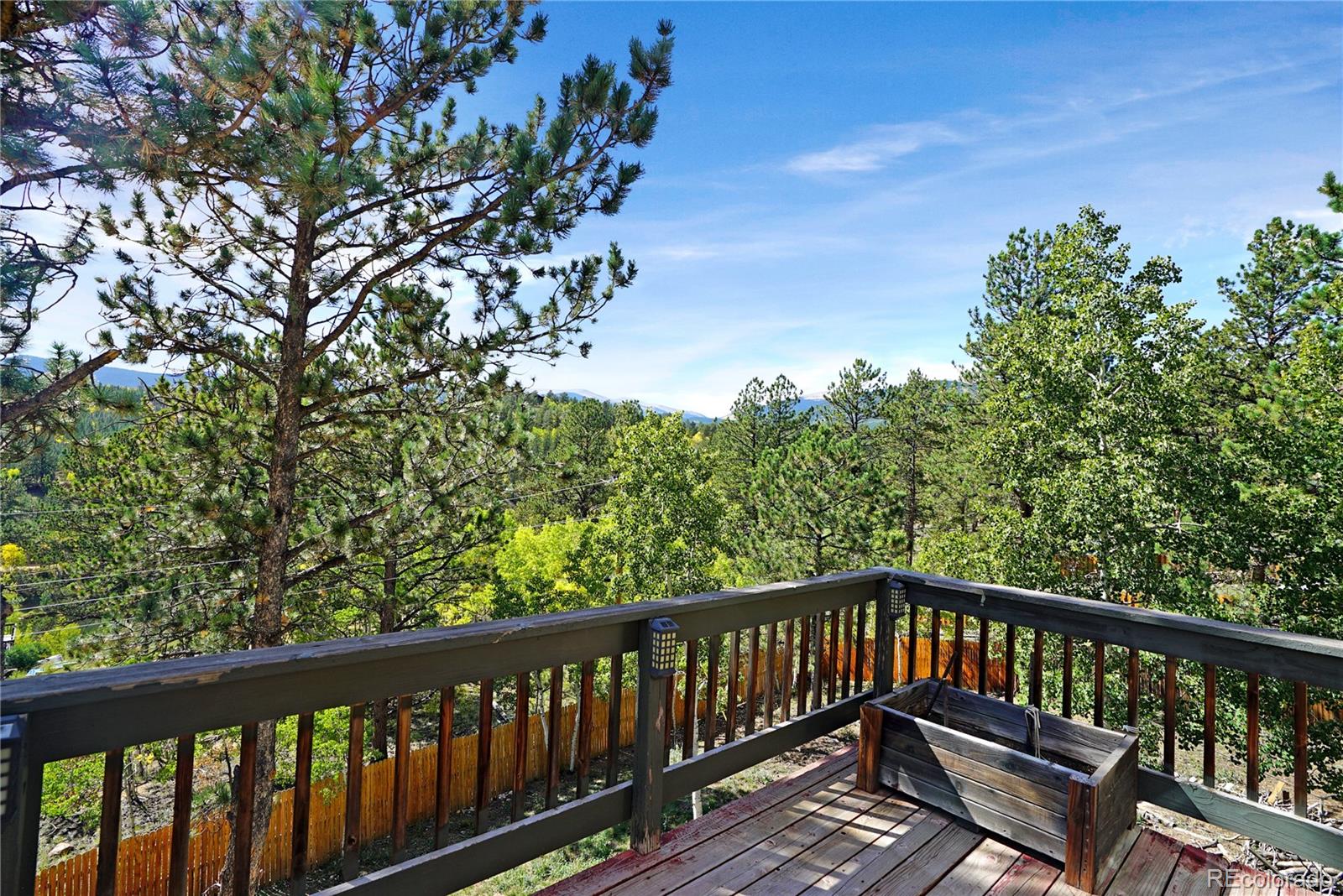 Wooden deck overlooking pine forest and distant mountain ridges beneath a blue sky.