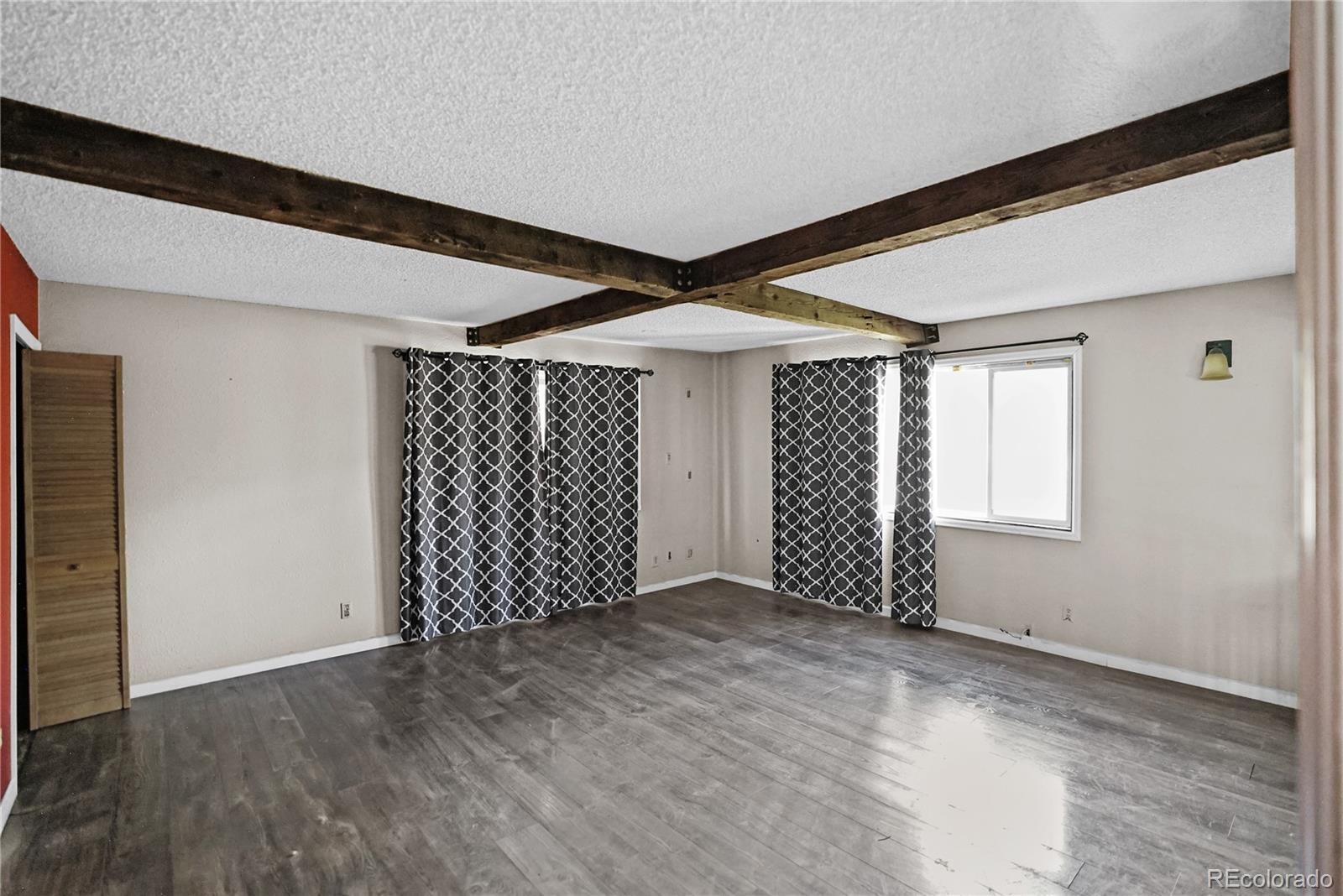 Empty living room with exposed ceiling beams, gray plank flooring, and patterned curtains.