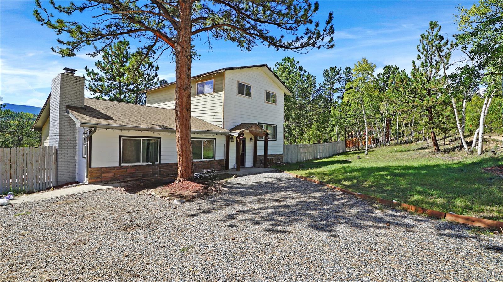 Two-story house with a brick chimney and gravel driveway among pine and aspen trees.
