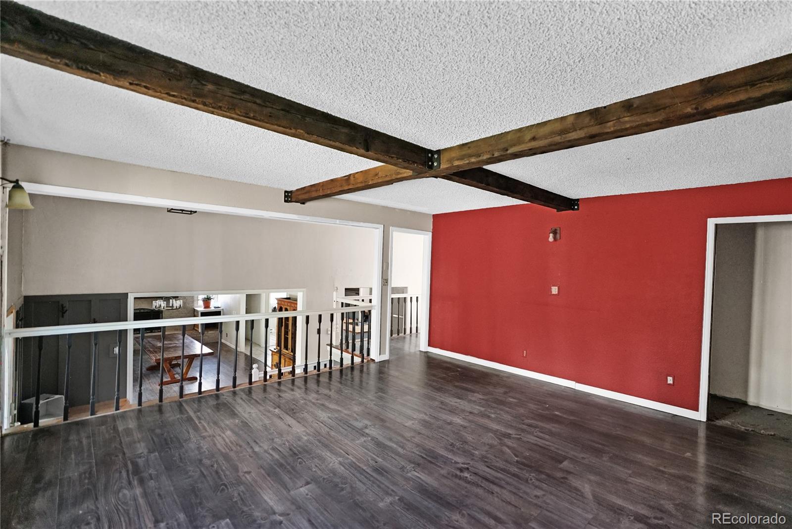 Upper-level loft with exposed timber beams, gray plank flooring, and red accent wall overlooking a furnished room.