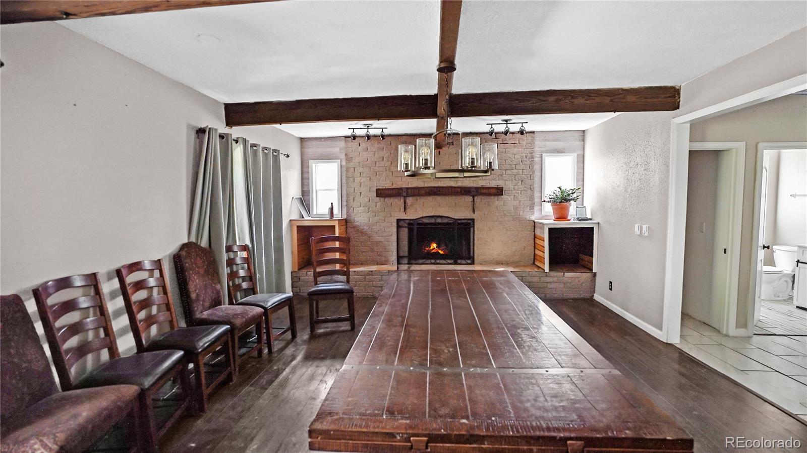 Dining area with a long wooden table, chairs, exposed beams, and a brick fireplace.