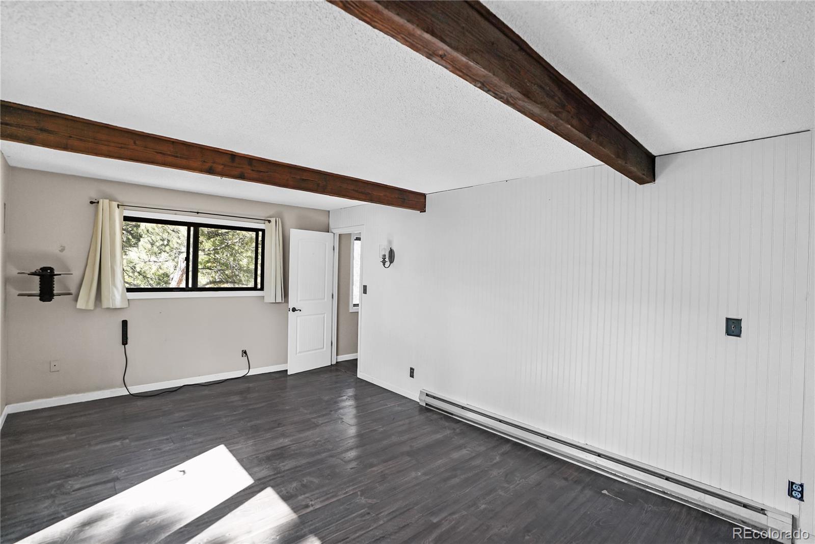 Vacant room with exposed wood ceiling beams, dark plank flooring, and a curtained window.