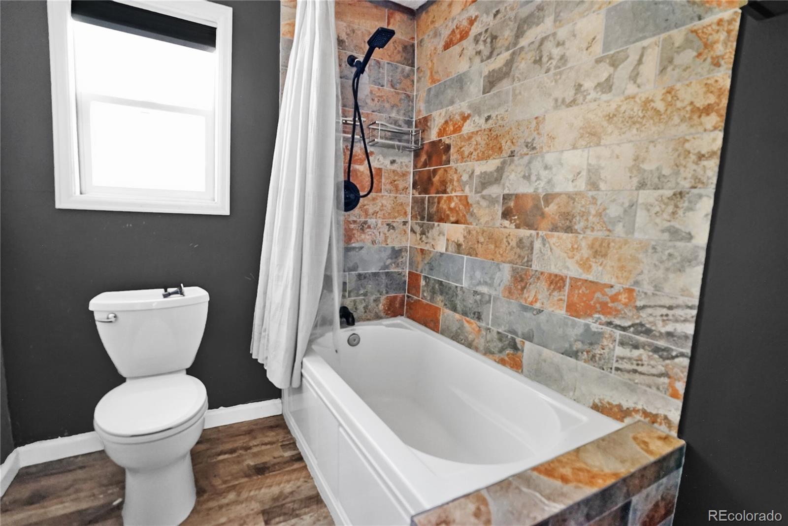 Bathroom with a white tub, multicolored tile surround, black shower fixtures, and a toilet.
