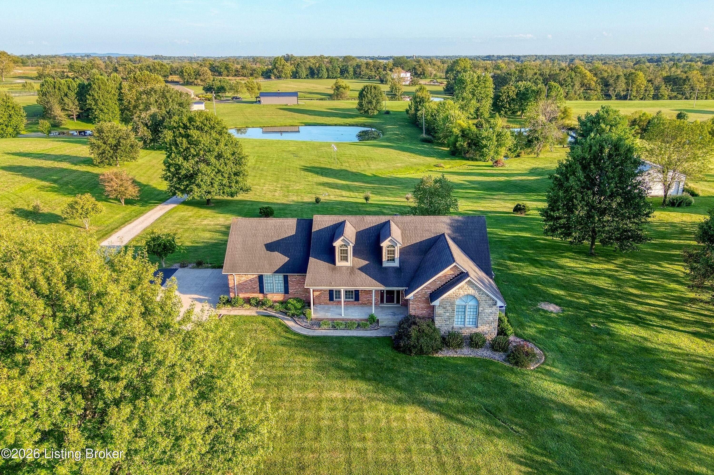 Aerial view of a brick and stone house on expansive green grounds, with a pond and mature trees.