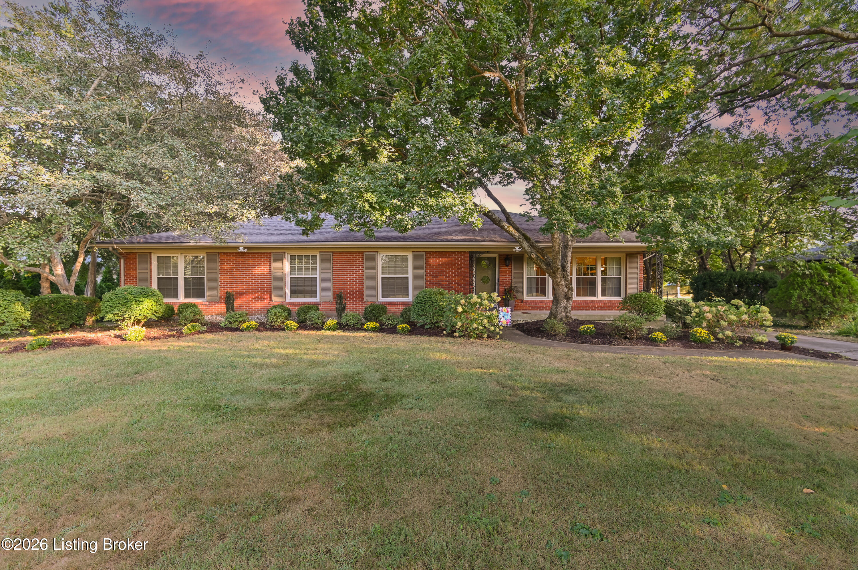 Red brick ranch house framed by mature trees, with landscaped shrubs and a broad front lawn.
