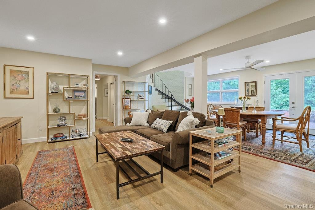 Open-plan living and dining room with a brown sectional, wood tables, staircase, and leafy views through large windows.