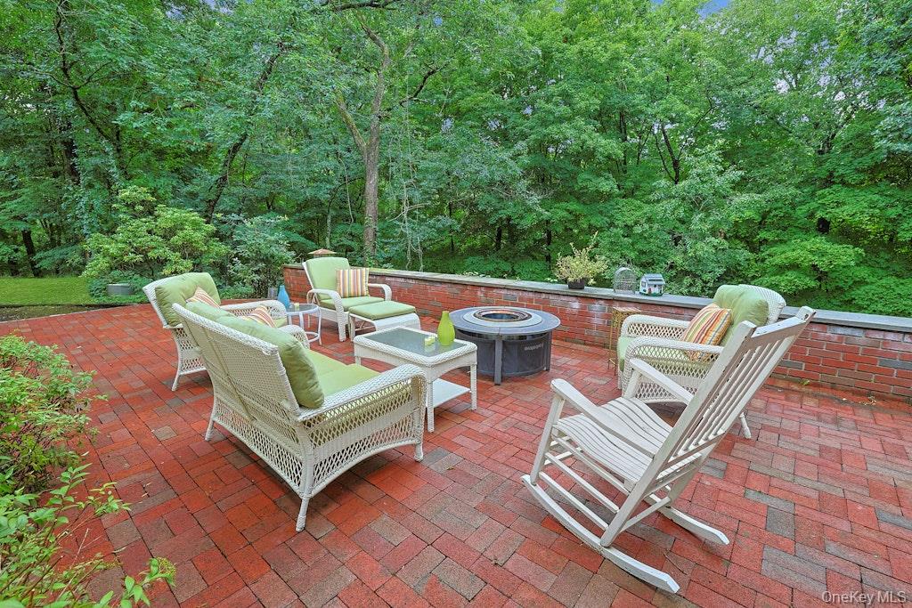 Furnished brick patio with wicker seating and a central fire table, surrounded by leafy woods.