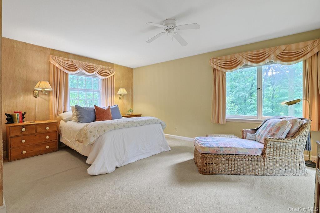 Furnished bedroom with a white bed and wicker seating beside large windows overlooking trees.