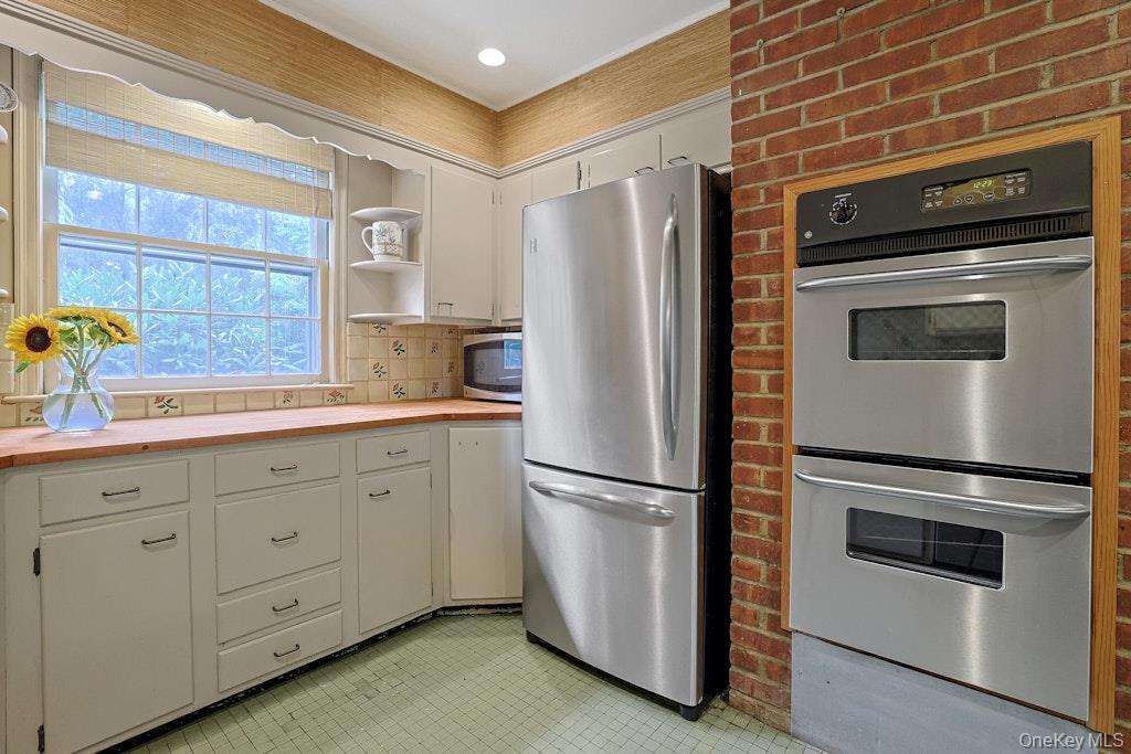 Kitchen with white cabinets, wood countertops, stainless refrigerator, and double oven set into a brick wall.