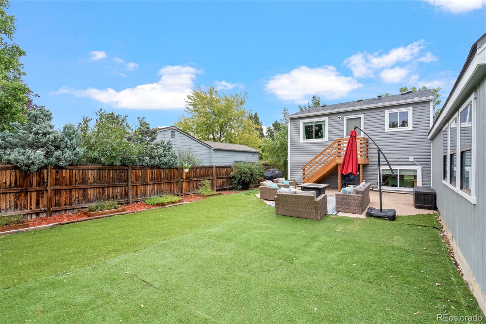 Fenced backyard with a broad lawn, patio seating, red umbrella, and wooden stairs to an elevated deck.