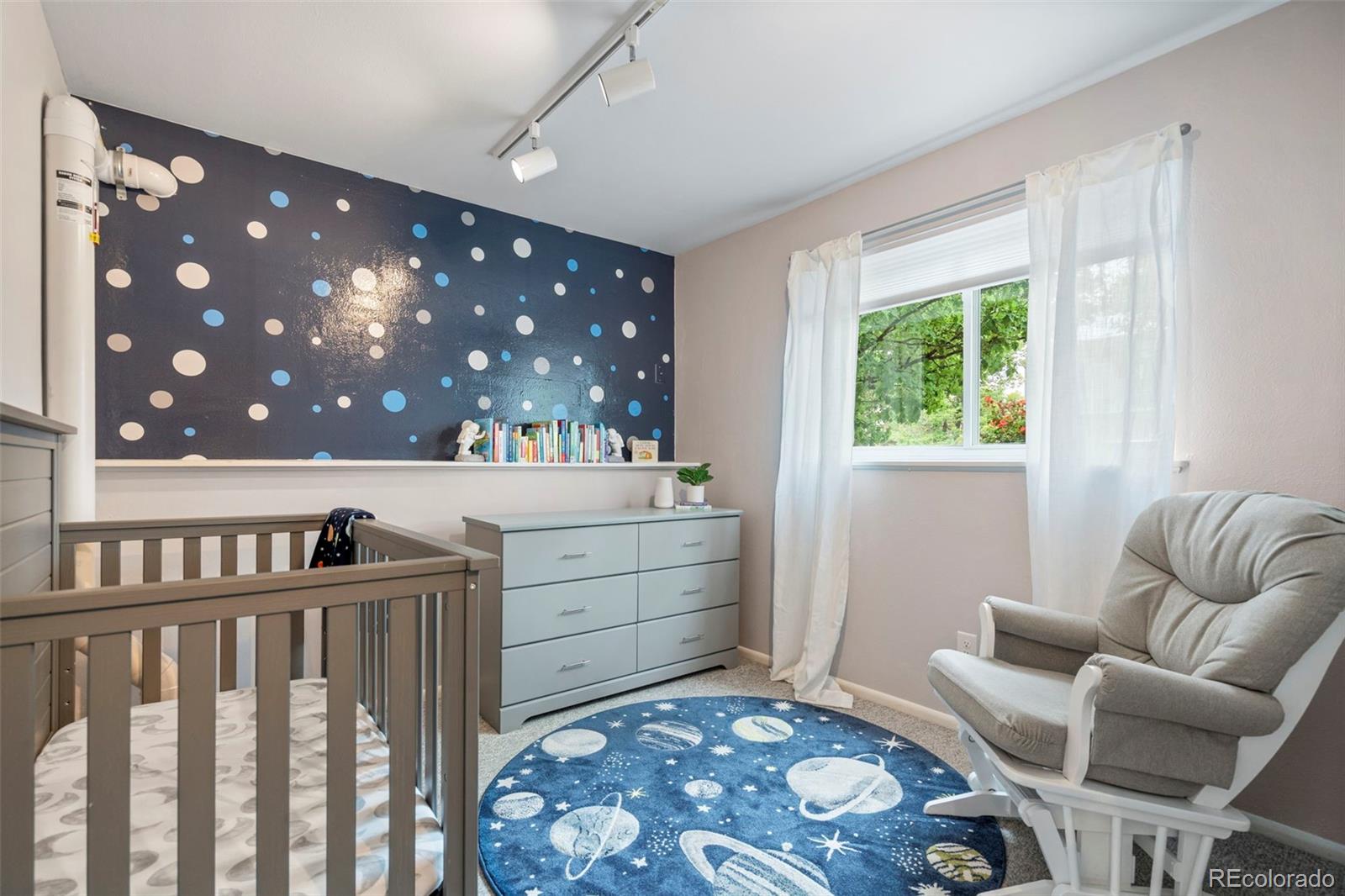 Nursery with a gray crib, space-themed rug, dotted accent wall, and cushioned rocking chair.