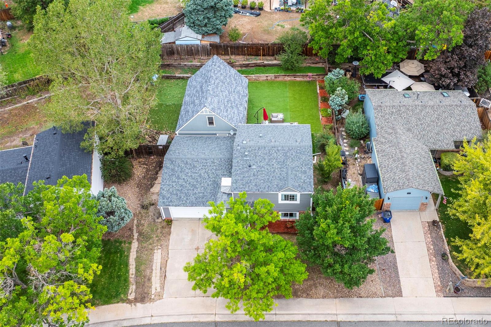 Aerial view of a gray-roofed home with a fenced green backyard and mature trees.