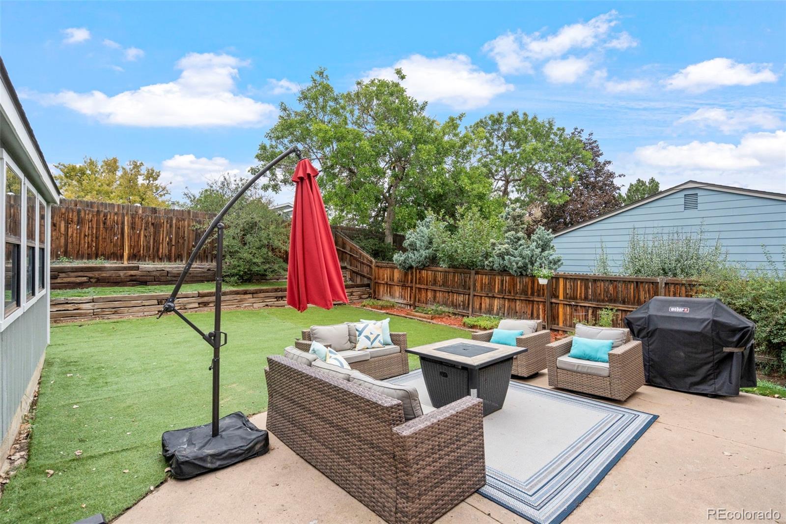 Backyard patio with wicker seating, a fire table, red umbrella, and artificial turf.