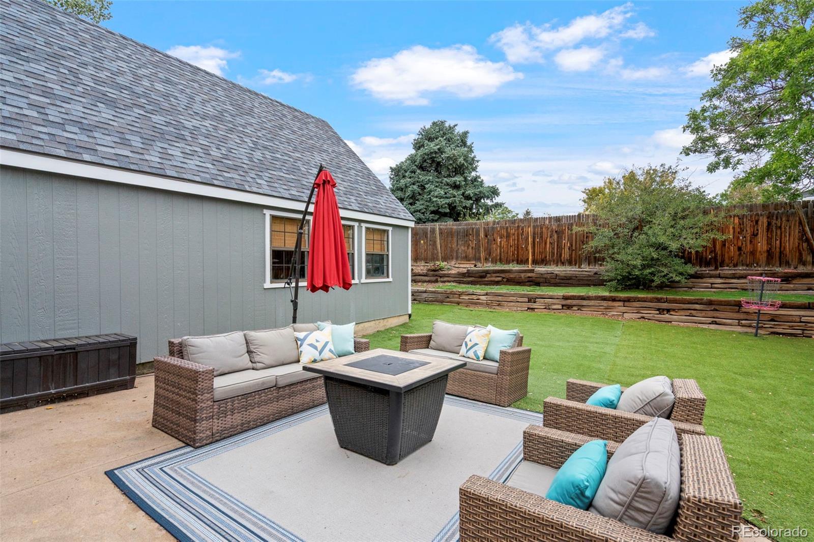 Backyard patio with wicker seating and a fire table beside a fenced lawn.