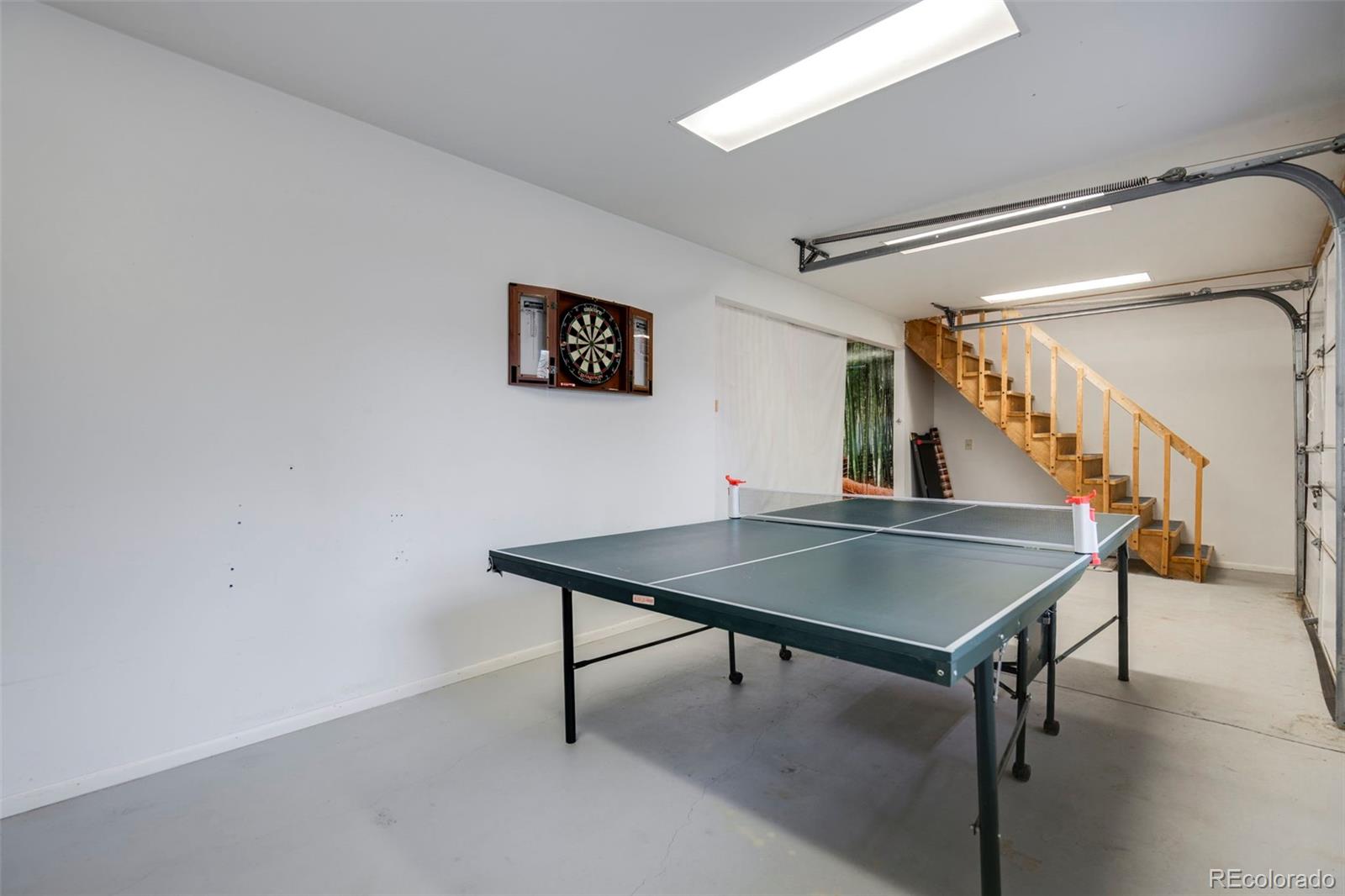 Garage game area with a table tennis table, wall-mounted dartboard, and wooden stairs.