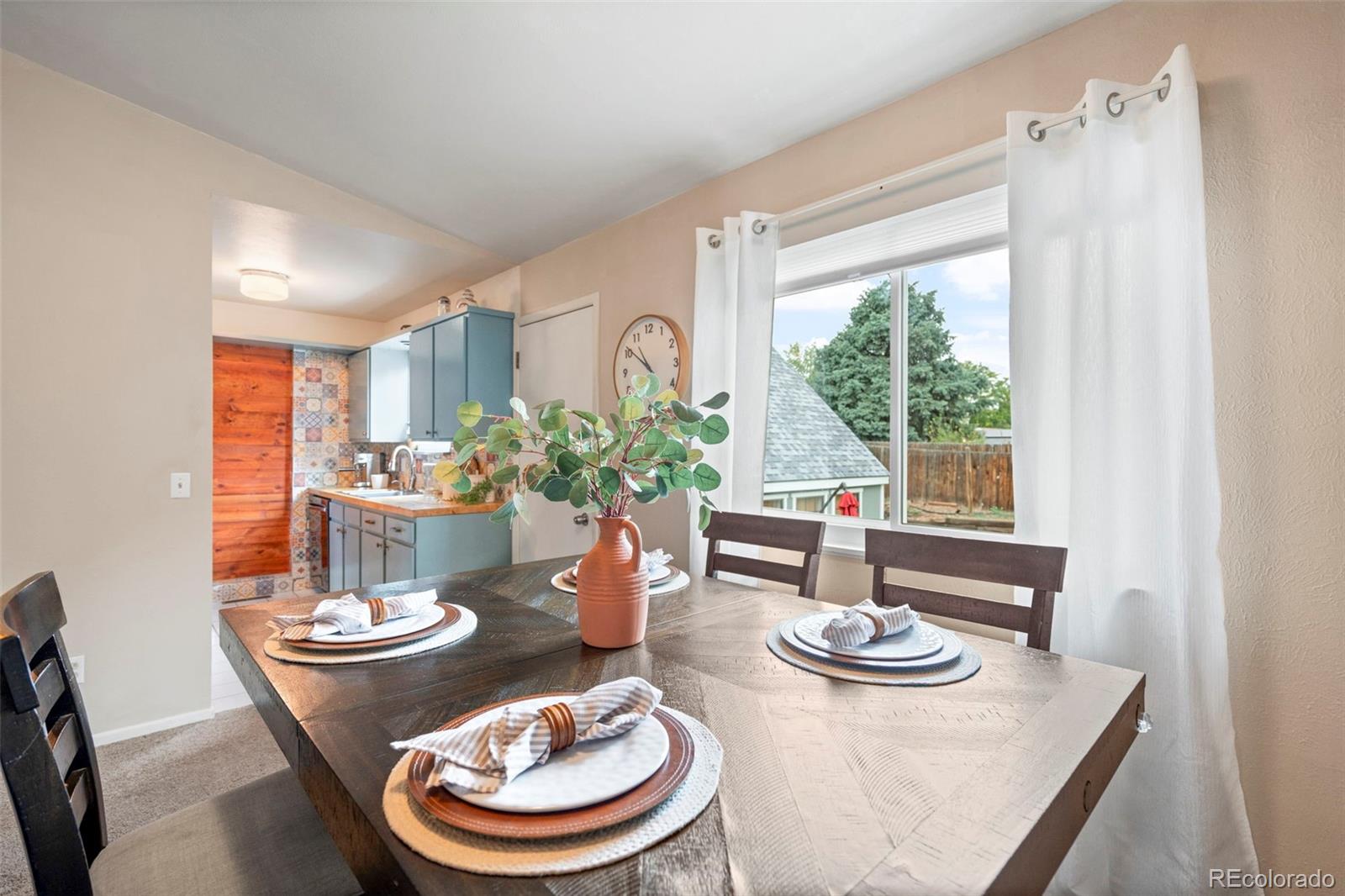 Dining area with a set table beside a large window, adjoining a kitchen with blue cabinets.