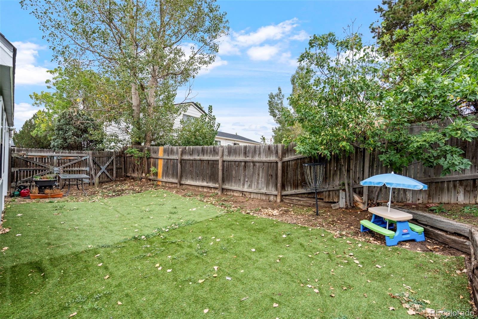 Fenced backyard with a green lawn, mature trees, patio seating, and a children's play table.