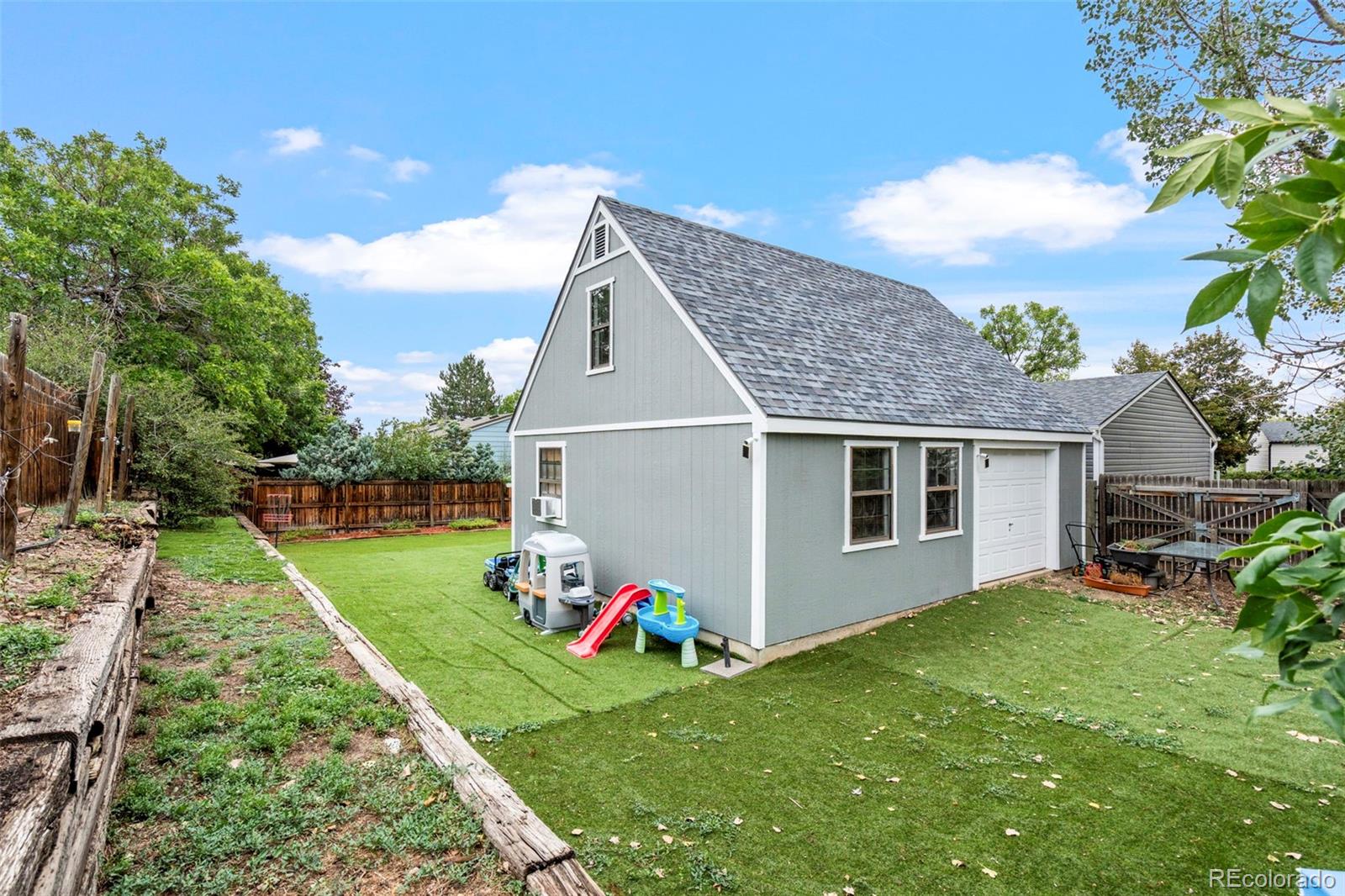 Gray barn-style garage in a fenced backyard with a broad lawn and raised garden beds.