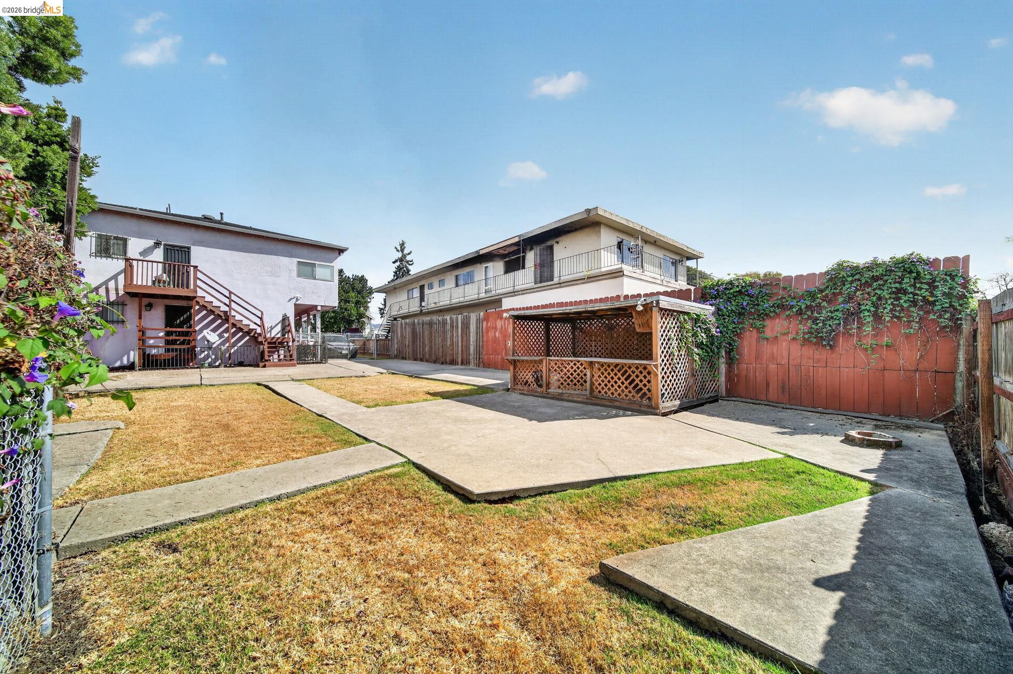 Fenced backyard with concrete patio paths, dry lawn, and a lattice gazebo beside a two-story building.