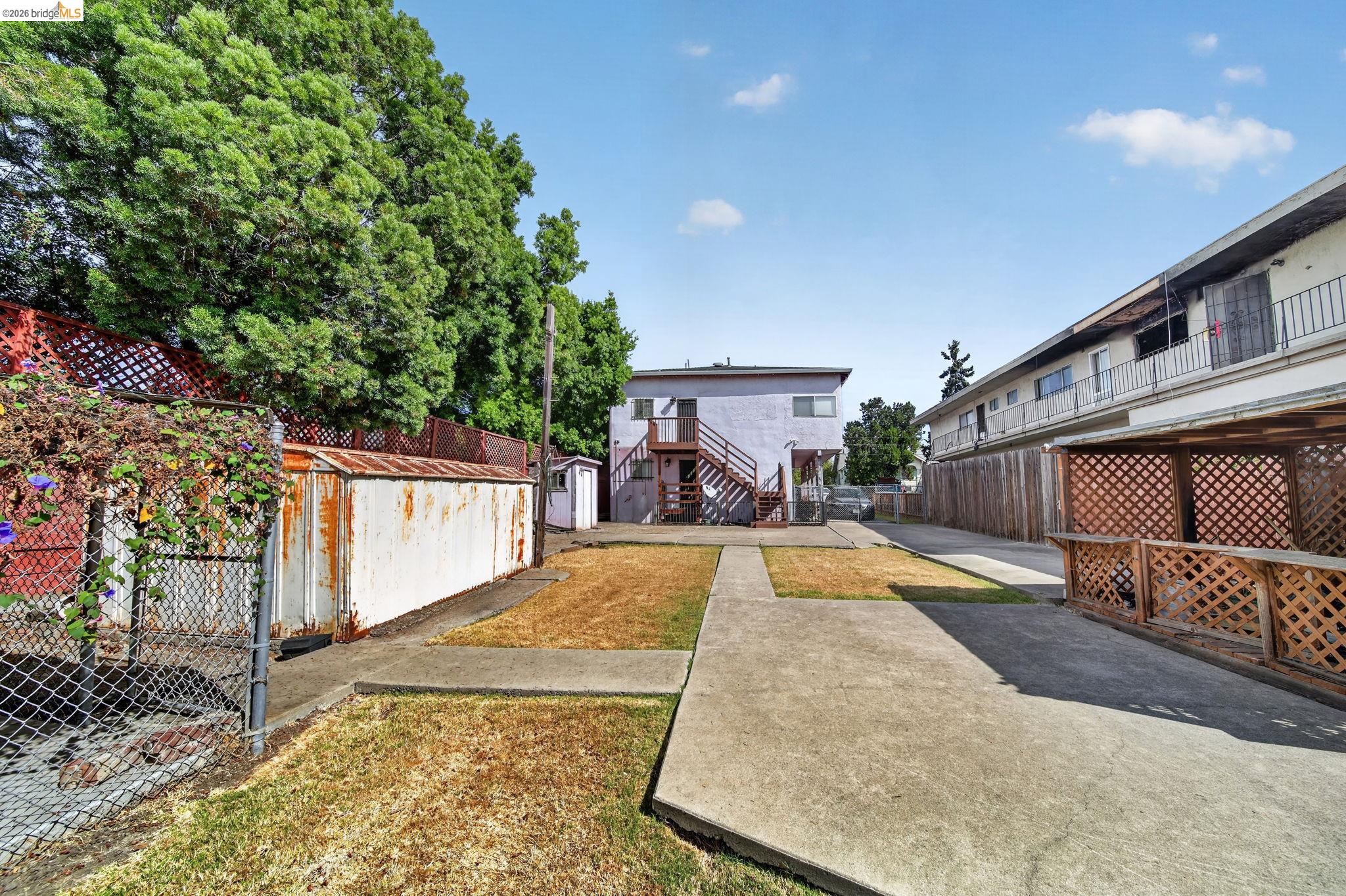 Shared courtyard between multi-unit buildings with concrete walkways, exterior stairs, lattice fencing, and mature trees.
