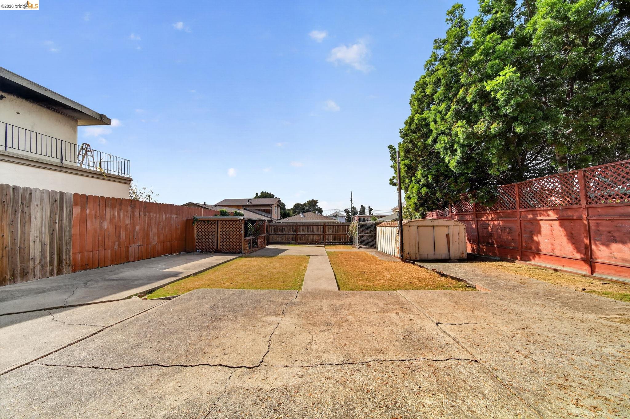 Fenced backyard with a cracked concrete patio, narrow lawn, central walkway, and small shed.