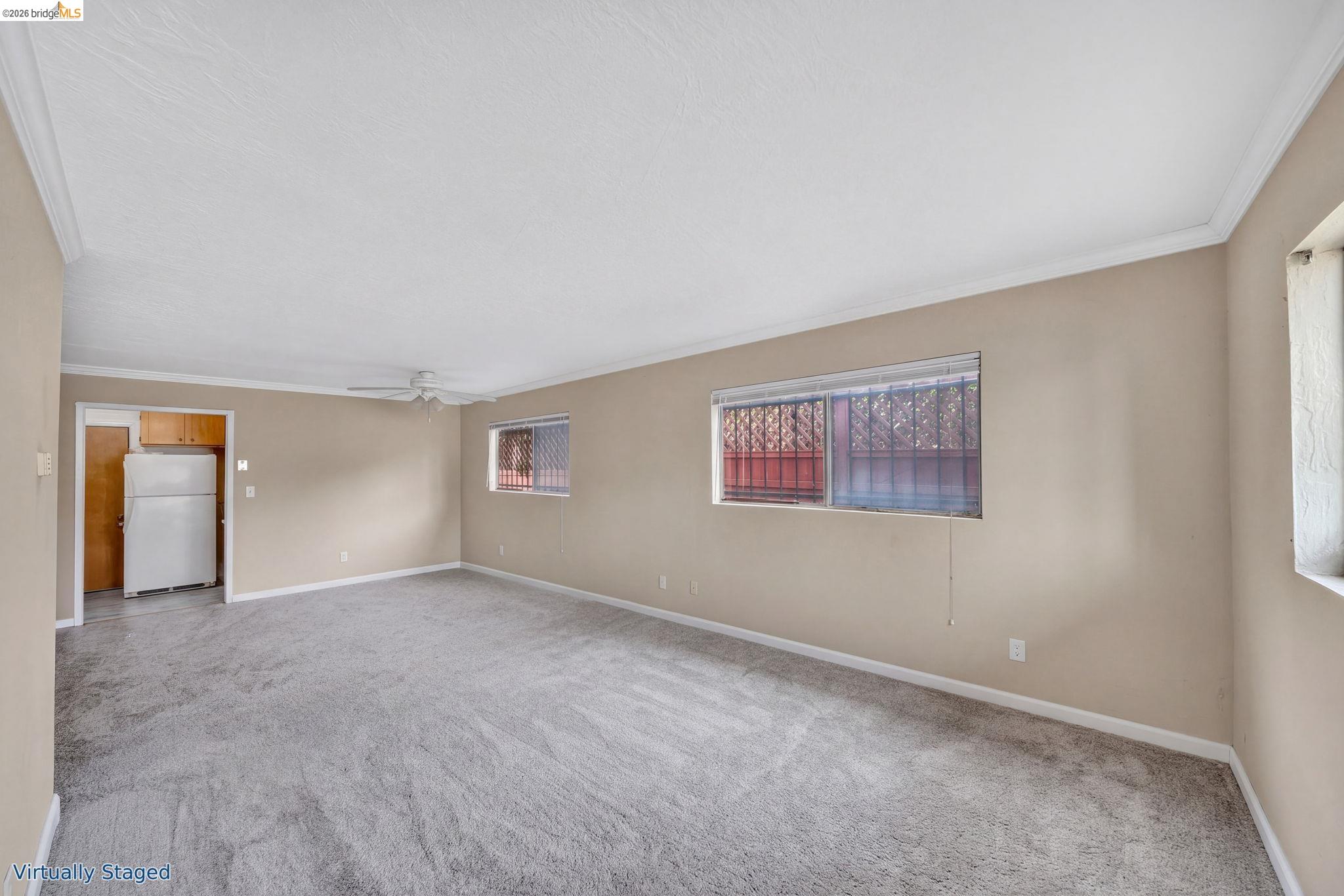 Empty living room with gray carpet, beige walls, ceiling fan, and barred windows.