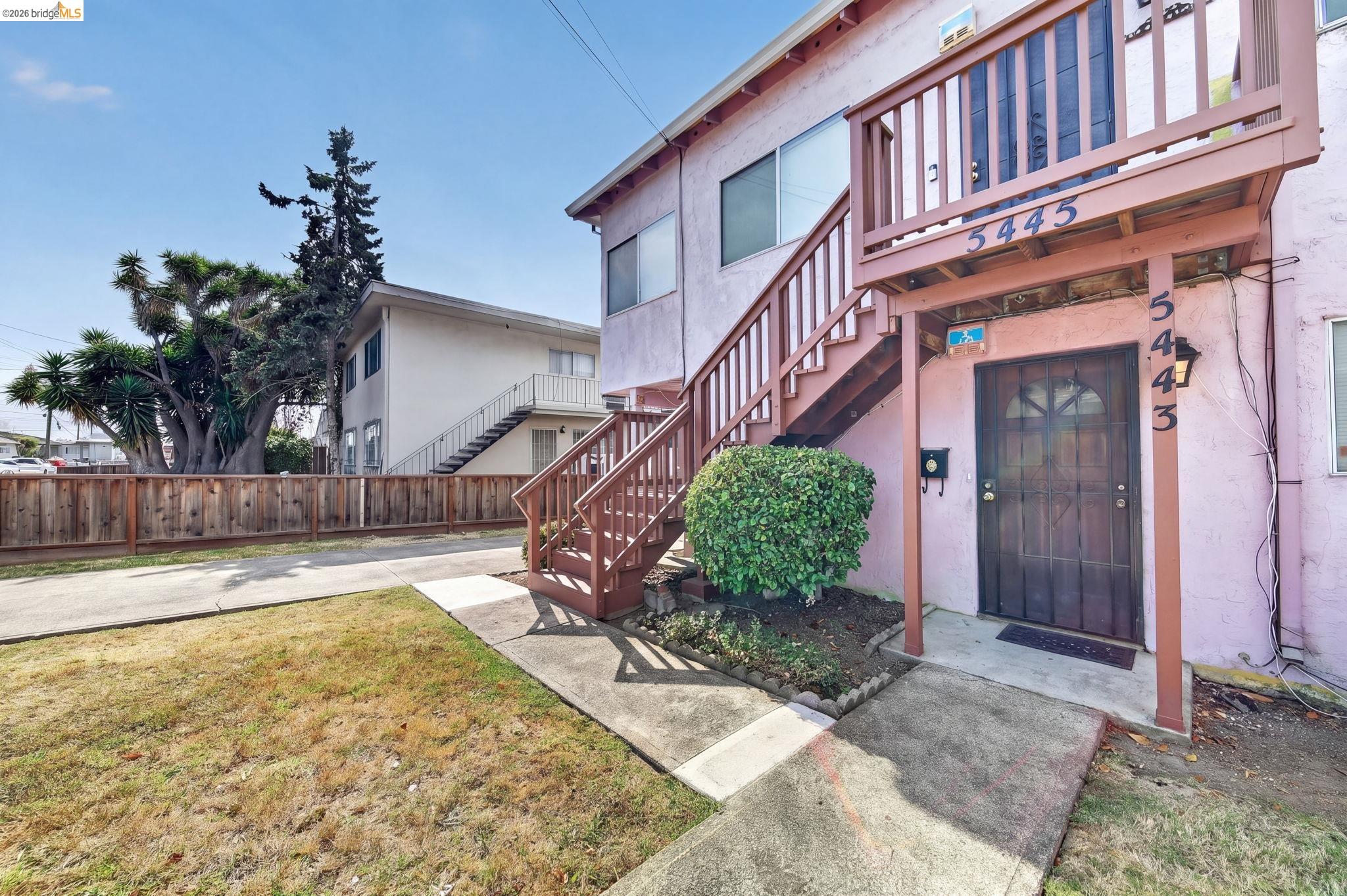 Pink building entrance with a dark door, raised wooden staircase, and small front lawn.
